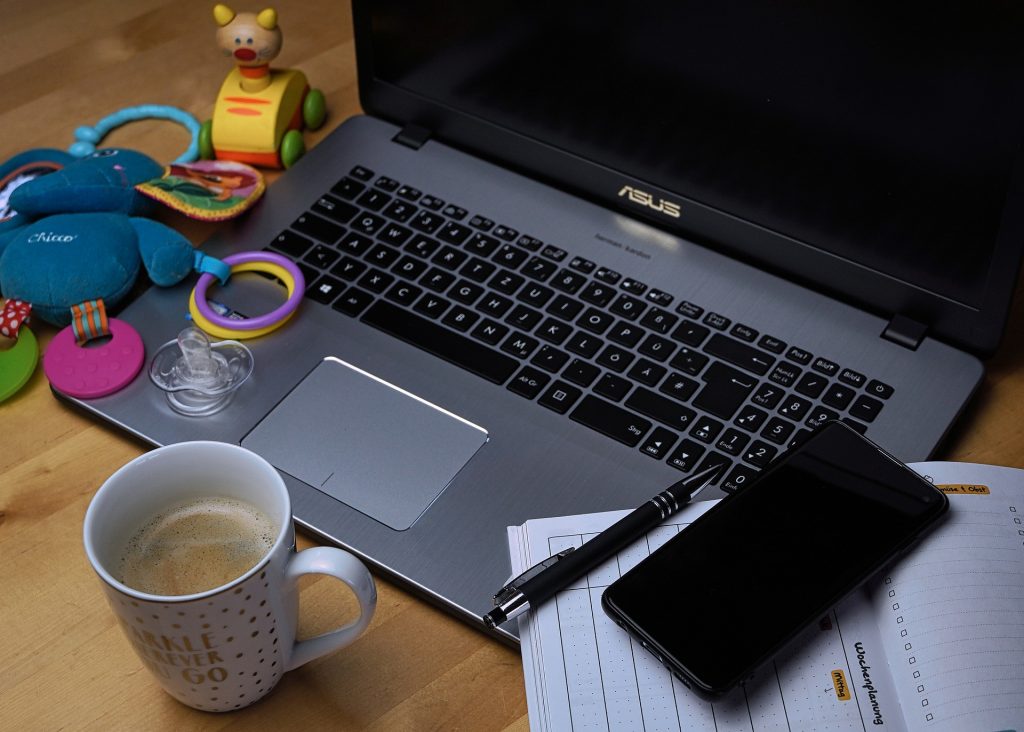 A laptop surrounded by coffee, a pad of paper, a cell phone and children toys., showing the struggle of working from home